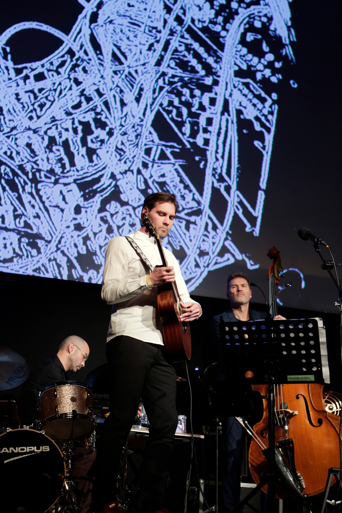 Auke Hulst draagt de tekst voor en speelt akoestische gitaar. Links achter hem Yonga Sun, rechts Hein Offermans. Foto Henk Veenstra